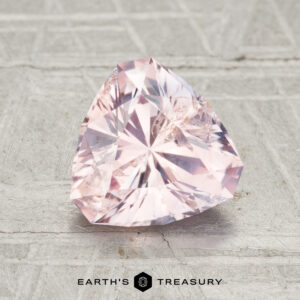 A 1.84-carat, faceted light pink Montana sapphire with a triangular shape rests on a textured gray surface, with "EARTH'S TREASURY" shown below.