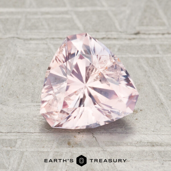 1.84-Carat Light Peach Pink Montana Sapphire A 1.84-carat, faceted light pink Montana sapphire with a triangular shape rests on a textured gray surface, with "EARTH'S TREASURY" shown below.