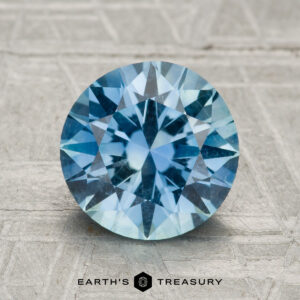 A 2.72-carat round brilliant-cut Montana sapphire is displayed on a textured gray surface, with "EARTH'S TREASURY" written below.