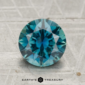 A 2.33-carat round brilliant-cut Montana sapphire is displayed on a textured gray surface, with the "Earth's Treasury" logo and text beneath it.