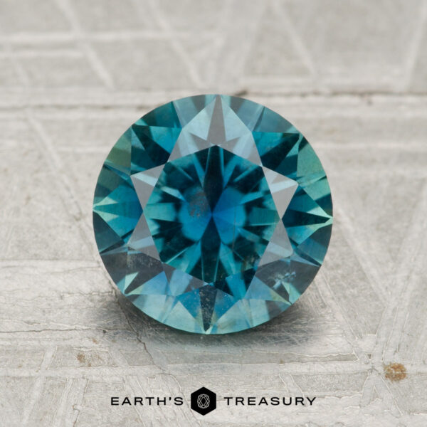 2.33-Carat Rich Teal Montana Sapphire A 2.33-carat round brilliant-cut Montana sapphire is displayed on a textured gray surface, with the "Earth's Treasury" logo and text beneath it.