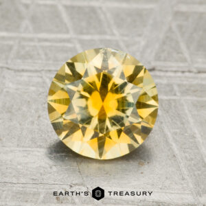 A 1.78-carat Montana Sapphire in a round brilliant cut is displayed on a textured gray surface, with the "Earth's Treasury" logo beneath it.