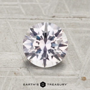 A 1.47-carat Montana sapphire, expertly cut in a round brilliant style, rests on a textured gray surface with "Earth's Treasury" displayed at the bottom.