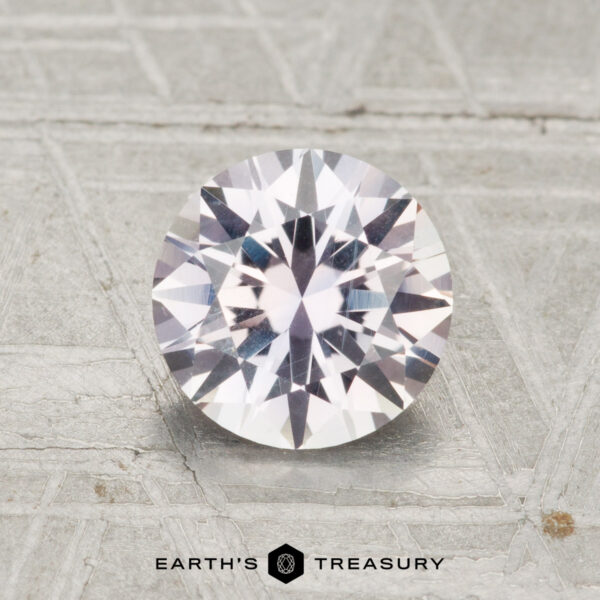 1.47-Carat Near-Colorless Montana Sapphire A 1.47-carat Montana sapphire, expertly cut in a round brilliant style, rests on a textured gray surface with "Earth's Treasury" displayed at the bottom.