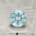 A 1.44-carat Montana sapphire, round and faceted with a light blue hue, rests on a textured grey surface featuring the "Earth's Treasury" branding at the bottom.