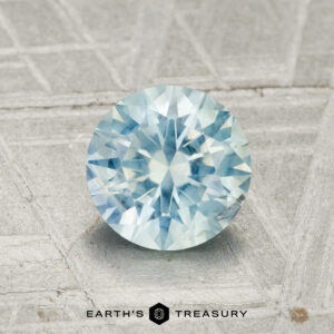 A 1.44-carat Montana sapphire, round and faceted with a light blue hue, rests on a textured grey surface featuring the "Earth's Treasury" branding at the bottom.