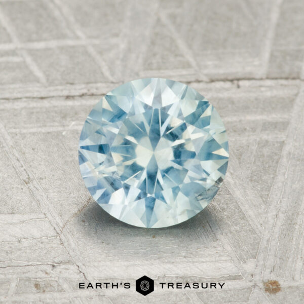 1.44-Carat Light Aqua Blue Montana Sapphire A 1.44-carat Montana sapphire, round and faceted with a light blue hue, rests on a textured grey surface featuring the "Earth's Treasury" branding at the bottom.