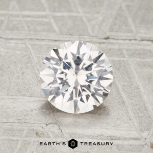A 1.30-carat Montana sapphire with a round brilliant cut rests on a textured gray surface, with "Earth's Treasury" visible at the bottom.