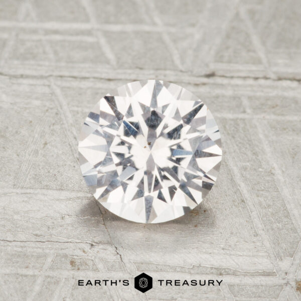 1.30-Carat Near-Colorless Montana Sapphire A 1.30-carat Montana sapphire with a round brilliant cut rests on a textured gray surface, with "Earth's Treasury" visible at the bottom.