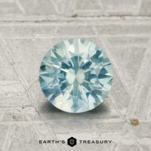 A 1.22-carat Montana Sapphire, round-cut and light blue, is shown on a textured gray surface with the Earth's Treasury logo at the bottom.