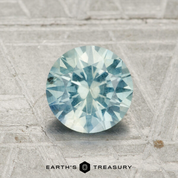 1.22-Carat Light Aqua Blue-Green Montana Sapphire A 1.22-carat Montana Sapphire, round-cut and light blue, is shown on a textured gray surface with the Earth's Treasury logo at the bottom.