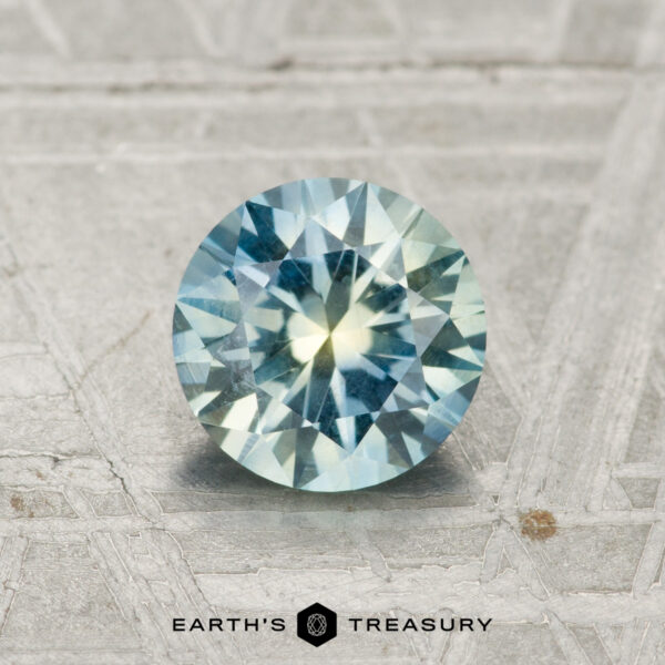 1.17-Carat Montana Sapphire A 1.17-carat Montana Sapphire, featuring round faceting and blue-yellow hues, rests on a textured metallic gray surface. "EARTH'S TREASURY" is displayed at the bottom.
