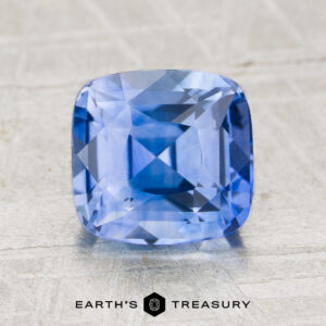 A 2.60-carat heated sapphire, cushion-cut and blue, is displayed on a textured surface with "EARTH'S TREASURY" and a hexagonal logo beneath it.