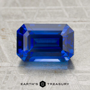 A 2.47-carat heated sapphire, emerald-cut and rectangular, is shown on a textured gray surface with the "EARTH'S TREASURY" logo at the bottom.