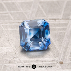 A faceted blue gemstone with an emerald cut, JV-001-795K, rests on a textured metallic surface. The "EARTH'S TREASURY" logo and text are displayed at the bottom of the image.