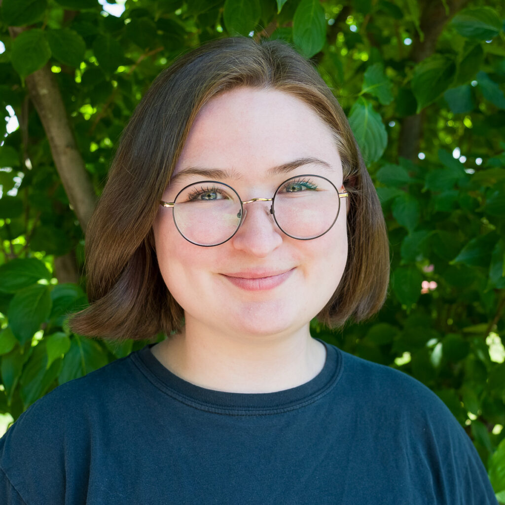 A person with short brown hair and round glasses smiles in front of leafy green foliage, wearing a black _DSC3610-1200-BW shirt.