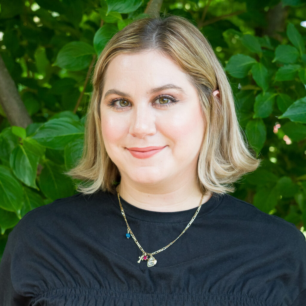 A woman with blonde hair in a black top stands outdoors before green bushes, smiling at the camera. Her natural beauty is captured in the photo titled _DSC4234-2-1200-BW.