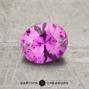 A 1.15-carat Pink Ceylon Sapphire (Heated), cut in a faceted oval shape, is displayed on a textured gray surface with the Earth’s Treasury logo beneath it.