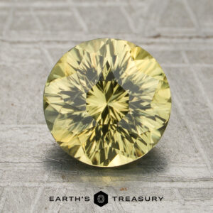 A 2.29-carat unheated greenish-yellow Kenya sapphire, round and faceted, is displayed upright on a gray patterned surface with the "Earth's Treasury" text and logo below it.