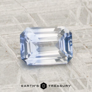 A rectangular, faceted 1.56-carat heated sapphire with a light blue hue is displayed on a textured gray surface, featuring the "Earth's Treasury" logo and text at the bottom.