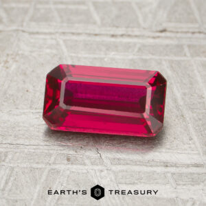 A 1.30-carat ruby, rectangular and faceted, is displayed on a textured gray surface. The text "EARTH'S TREASURY" appears at the bottom of the image.