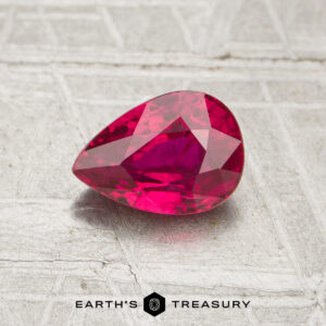A 1.07-carat, pear-shaped faceted ruby rests on a textured, light surface with the text "EARTH'S TREASURY" below.