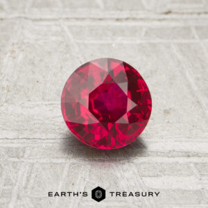 A 1.12-carat round, faceted ruby is displayed on a textured light gray surface. The "EARTH'S TREASURY" logo is shown at the bottom of the image.