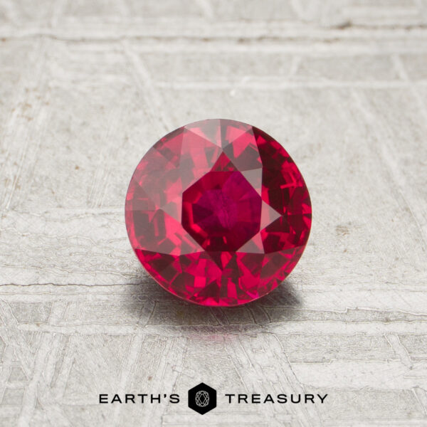 A 1.12-carat round, faceted ruby is displayed on a textured light gray surface. The "EARTH'S TREASURY" logo is shown at the bottom of the image.