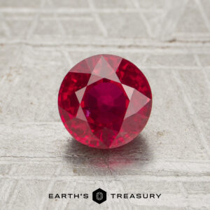 A 1.38-carat faceted round ruby is displayed on a textured surface with the text "EARTH'S TREASURY" beneath it.