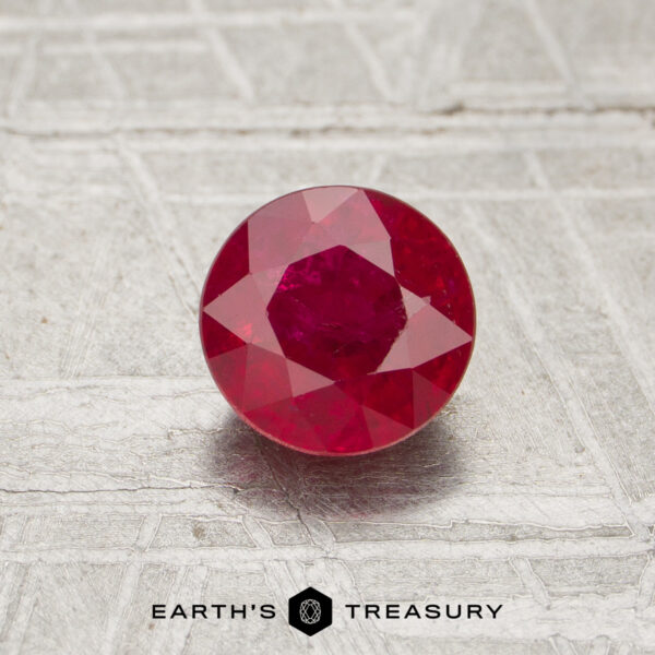 1.08-Carat Ruby A 1.08-carat round-cut ruby gemstone rests on a textured silver surface with the "EARTH'S TREASURY" logo and text at the bottom.