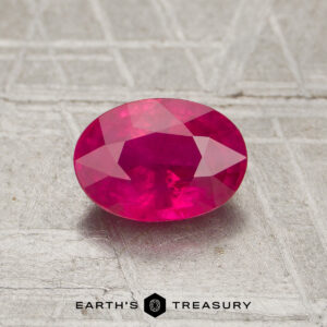 A 1.09-carat oval-cut vivid pink ruby rests on a textured gray surface, with the "Earth's Treasury" logo below it.