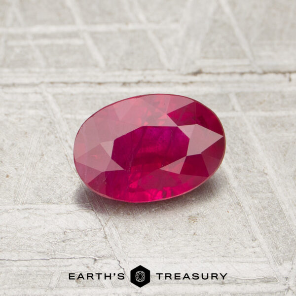 0.87-Carat Ruby A 0.87-carat faceted oval ruby rests on a textured light gray surface, with "EARTH'S TREASURY" and a hexagon logo displayed below the image.