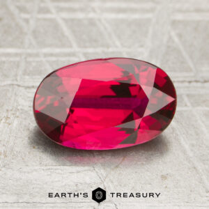 A 2.07-carat oval-cut deep red ruby is displayed on a textured surface, with "EARTH'S TREASURY" written below the gemstone.