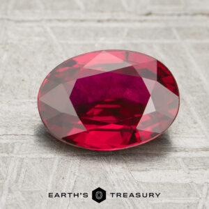 A 2.21-carat ruby with an oval faceted cut is displayed on a textured gray surface, with the text "EARTH'S TREASURY" below.
