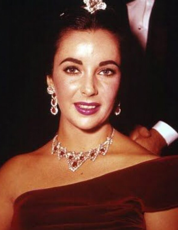 A woman with dark hair styled up wears a burgundy off-shoulder dress and the "cropped-The-Story-Behind-Elizabeth-Taylors-Iconic-Ruby-Necklace-1200-BW," posing against a dark background.