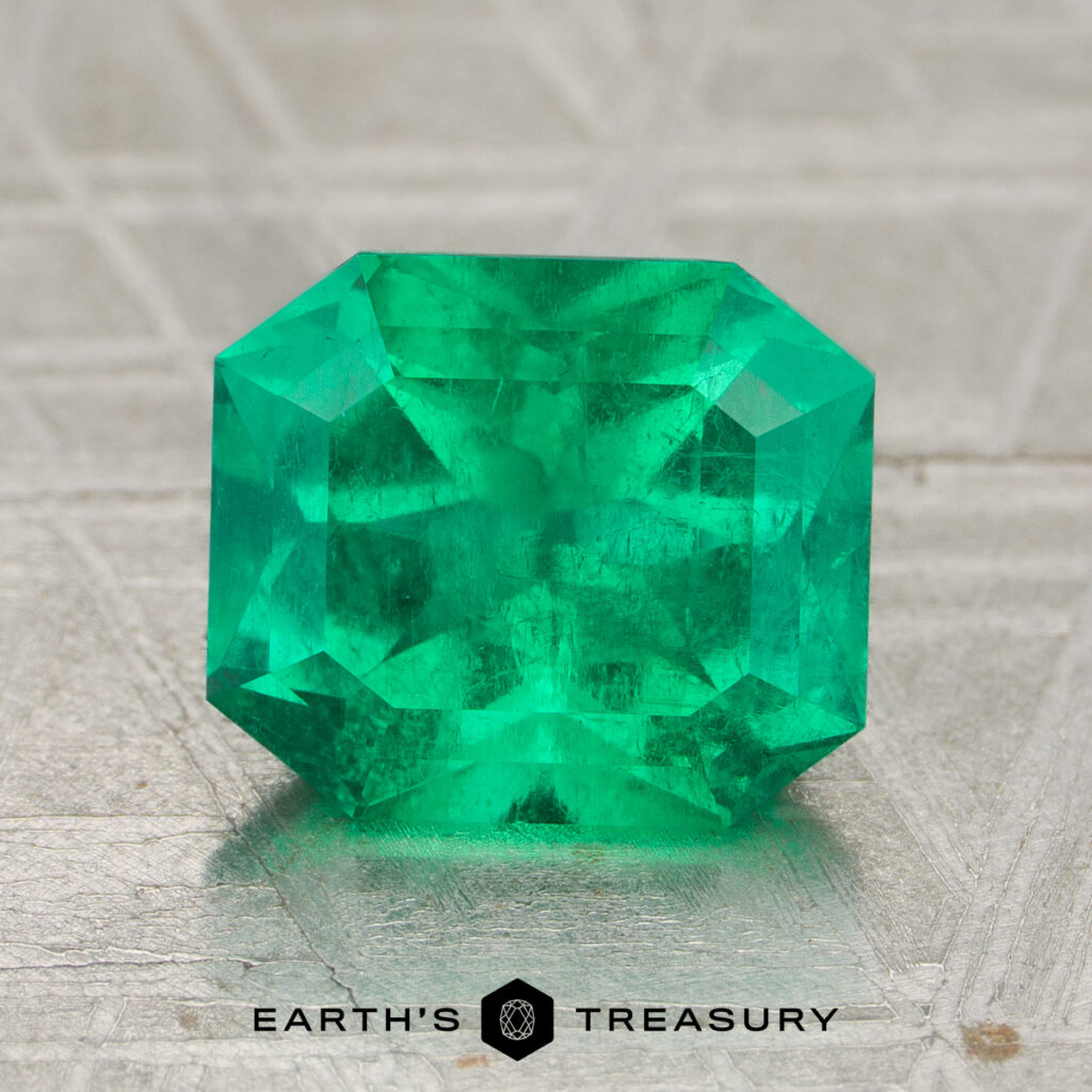 A 2.12-carat rich green Colombian emerald (oiled), cut in a rectangular faceted shape, displayed on a textured surface with the text "Earth's Treasury" below.