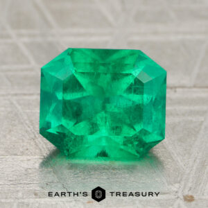 A 2.12-carat rich green Colombian emerald (oiled), cut in a rectangular faceted shape, displayed on a textured surface with the text "Earth's Treasury" below.