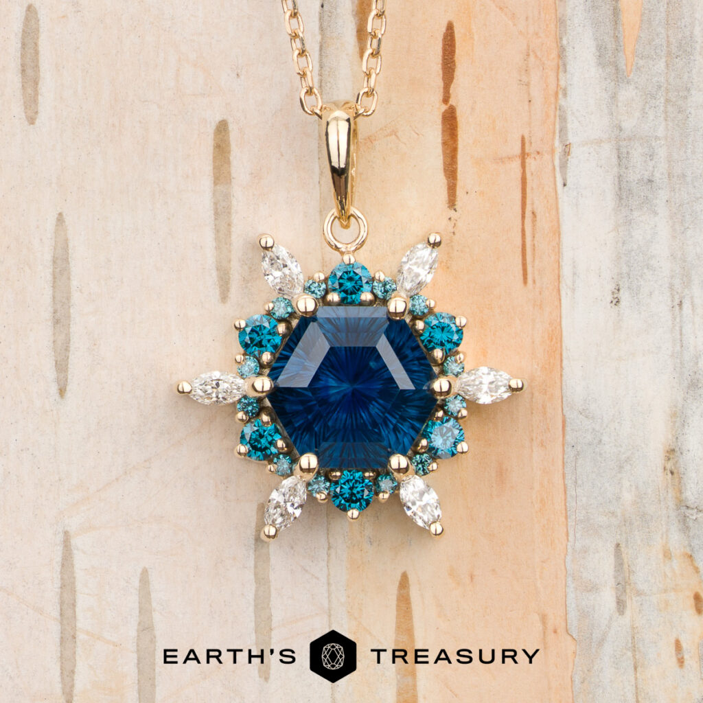 Gold necklace from the Lang, Michael [custom gallery], featuring a blue hexagonal gemstone pendant encircled by small blue stones and clear marquise gems, displayed on a wooden surface with the "Earth's Treasury" logo at the bottom.