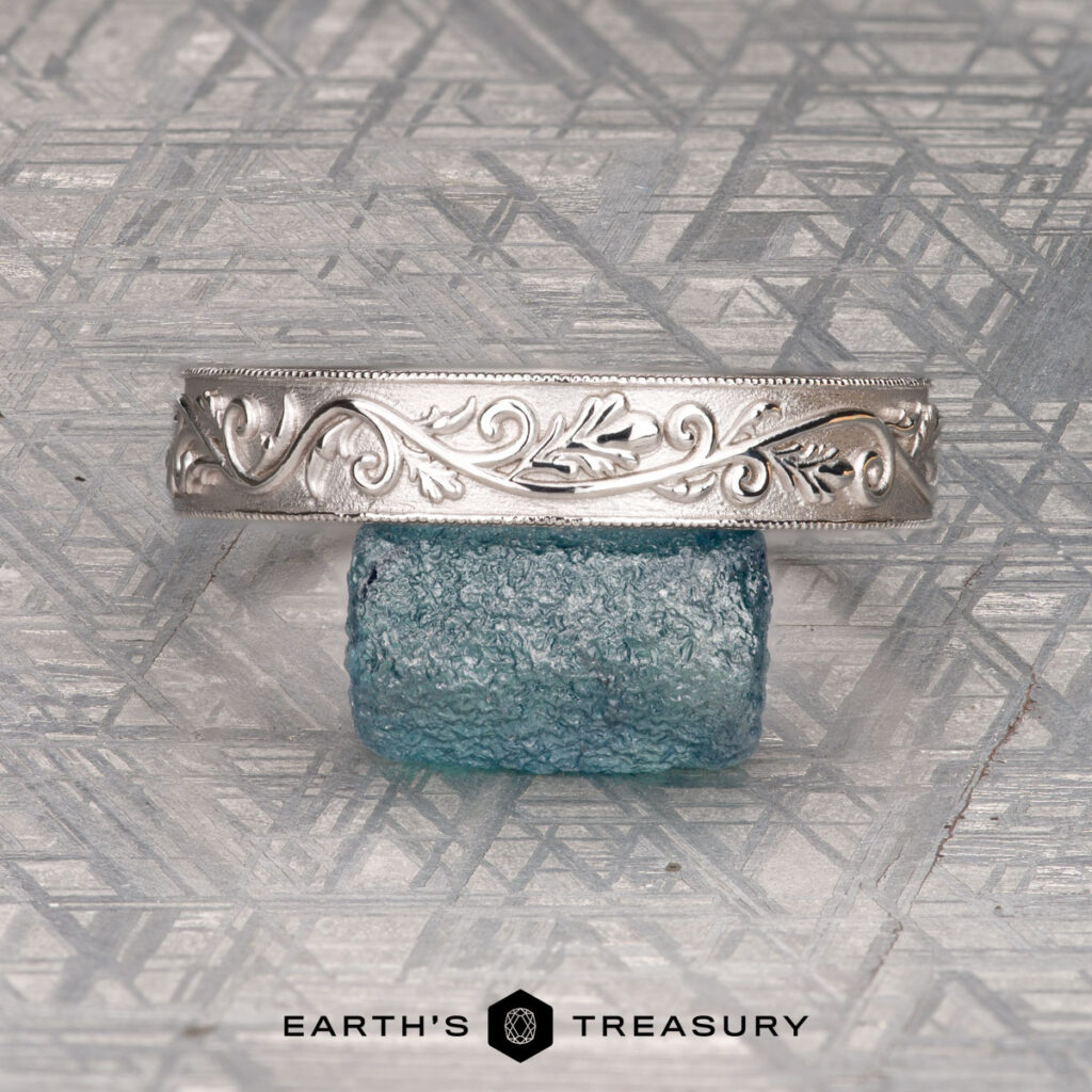A silver cuff bracelet, titled "Matt, Alexandr and Rondinelli, Megan 3," featuring an engraved leaf design and delicate filigree detailing, is displayed resting on a textured blue glass block against a patterned background. The text "EARTH'S TREASURY" appears at the bottom.