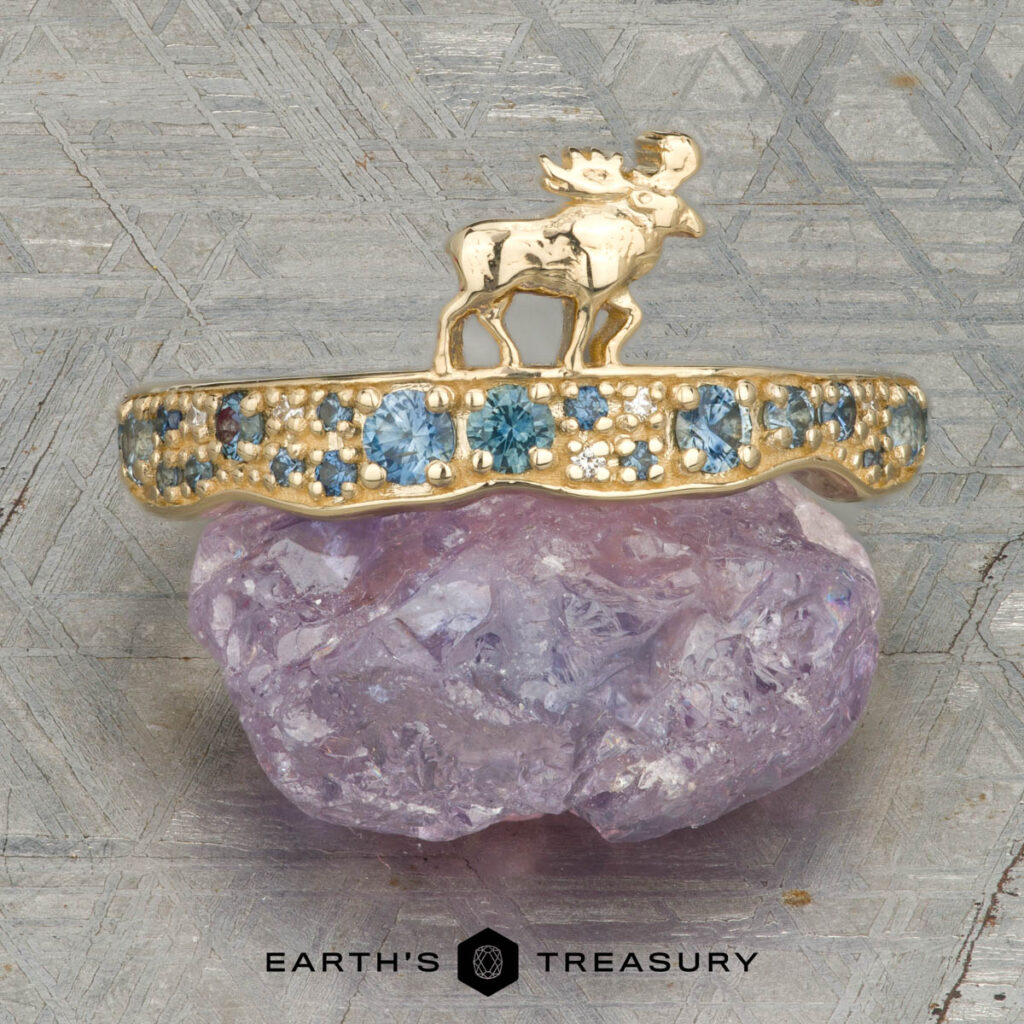 Blackall, Heather 2 [custom gallery]—a gold ring featuring a moose design and blue gemstones, displayed on a rough purple crystal with the "Earth’s Treasury" logo beneath.