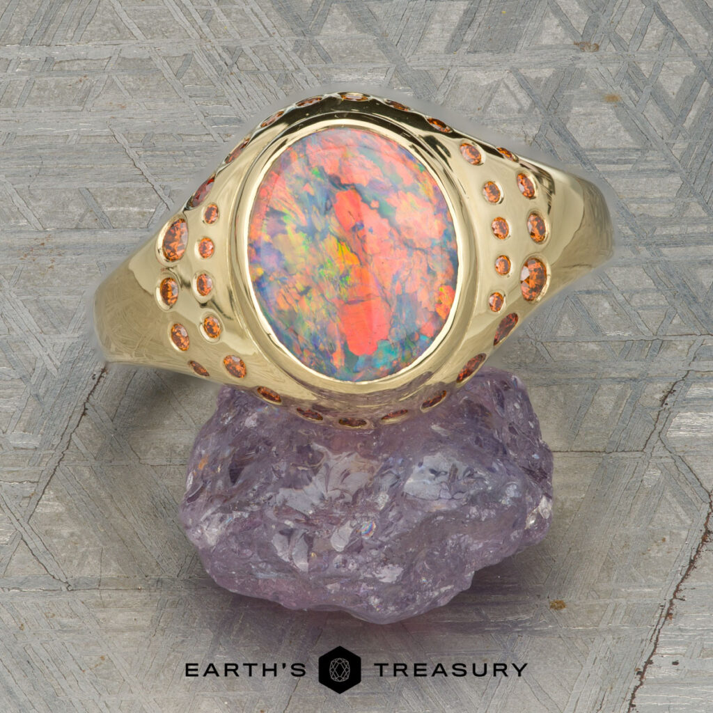 Chalchinsky, Jeremy [custom gallery] gold ring featuring an oval opal center stone and small round accent stones, displayed on a purple crystal with a geometric patterned background.