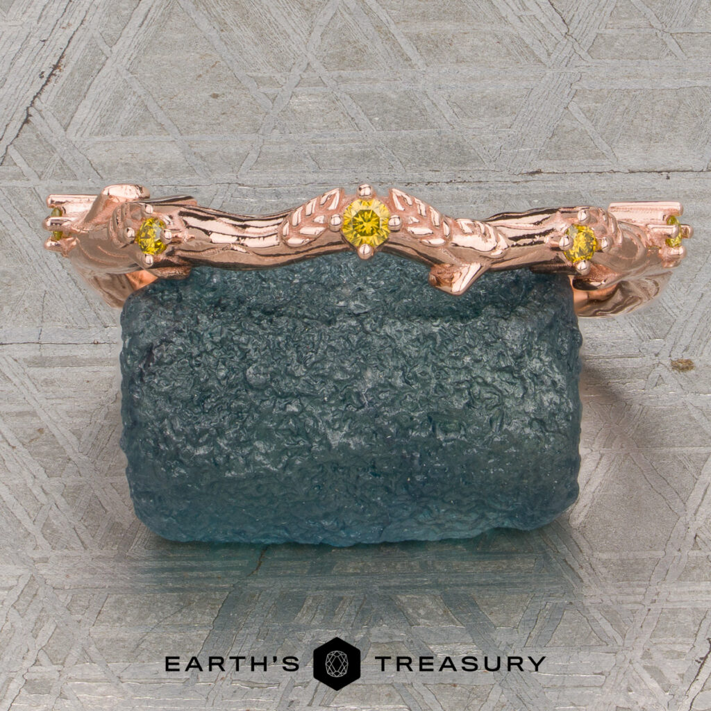 A rose gold ring, "Waldman, Jacob" from the custom gallery, featuring a branch design and yellow gemstones, is displayed on a rough blue stone. The Earth's Treasury logo appears at the bottom of the image.