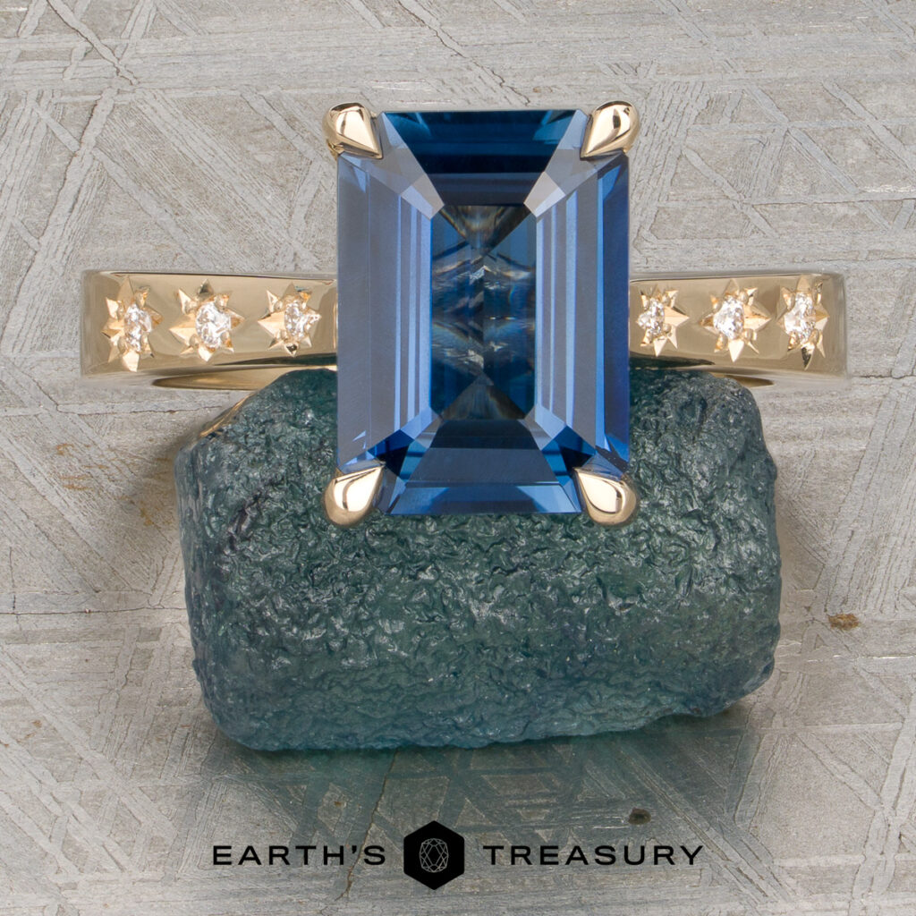 A custom gallery ring by Druger, Brayden and Langman, Becca features a rectangular blue gemstone accented by star-set diamonds on a gold band, displayed atop a rough blue stone. The "Earth's Treasury" logo and text are shown at the bottom.