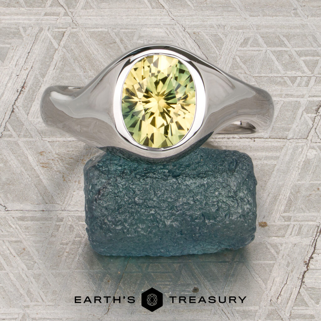 A silver ring with an oval, faceted yellow-green gemstone from the Kim, Eric [custom gallery] collection rests on a rough blue mineral. The background features a patterned gray surface, with the “Earth’s Treasury” logo and text displayed below.