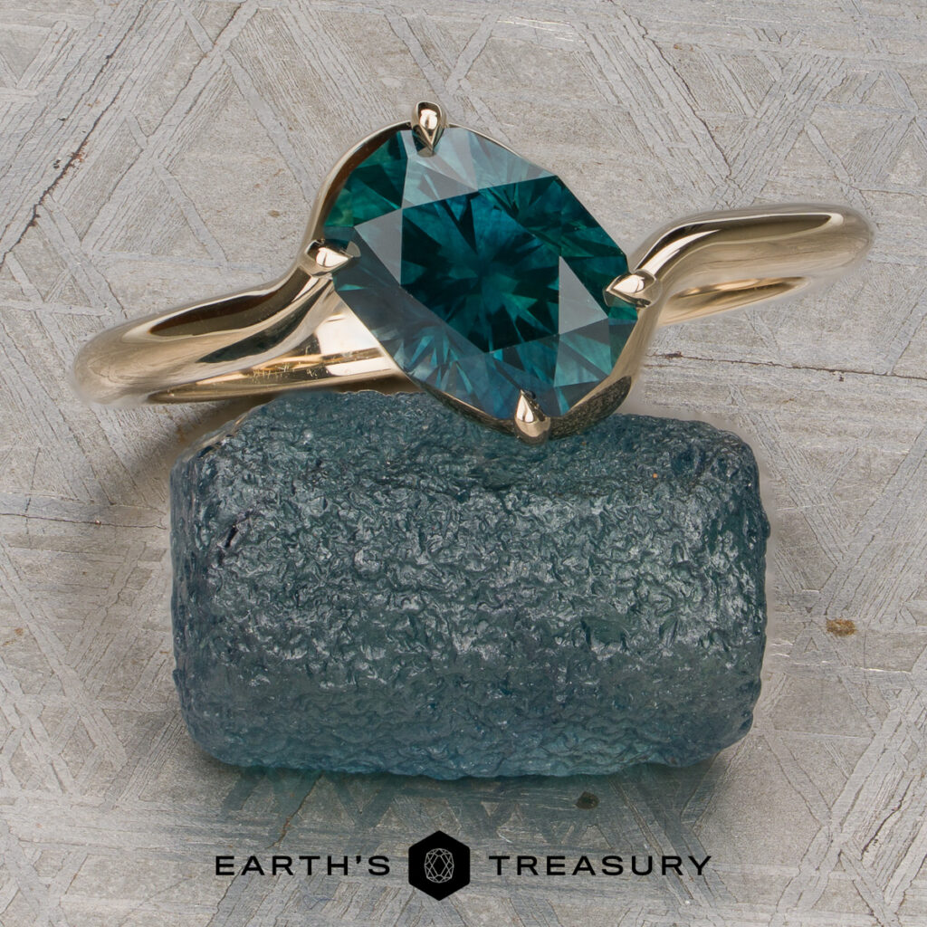 A custom-designed ring from Pitts, Jeremiah and Price, McCall [custom gallery], featuring a blue-green faceted gemstone set in gold, rests on a rough blue crystal atop a textured surface, with the "Earth's Treasury" logo beneath.