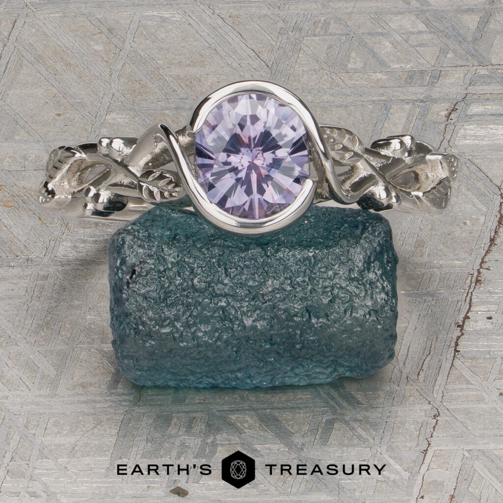 A silver ring from Sparks, Brad [custom gallery], featuring a round, pale purple gemstone and leaf-like designs on the band, rests on a rough blue-green stone. Below are the "EARTH'S TREASURY" name and hexagon logo.