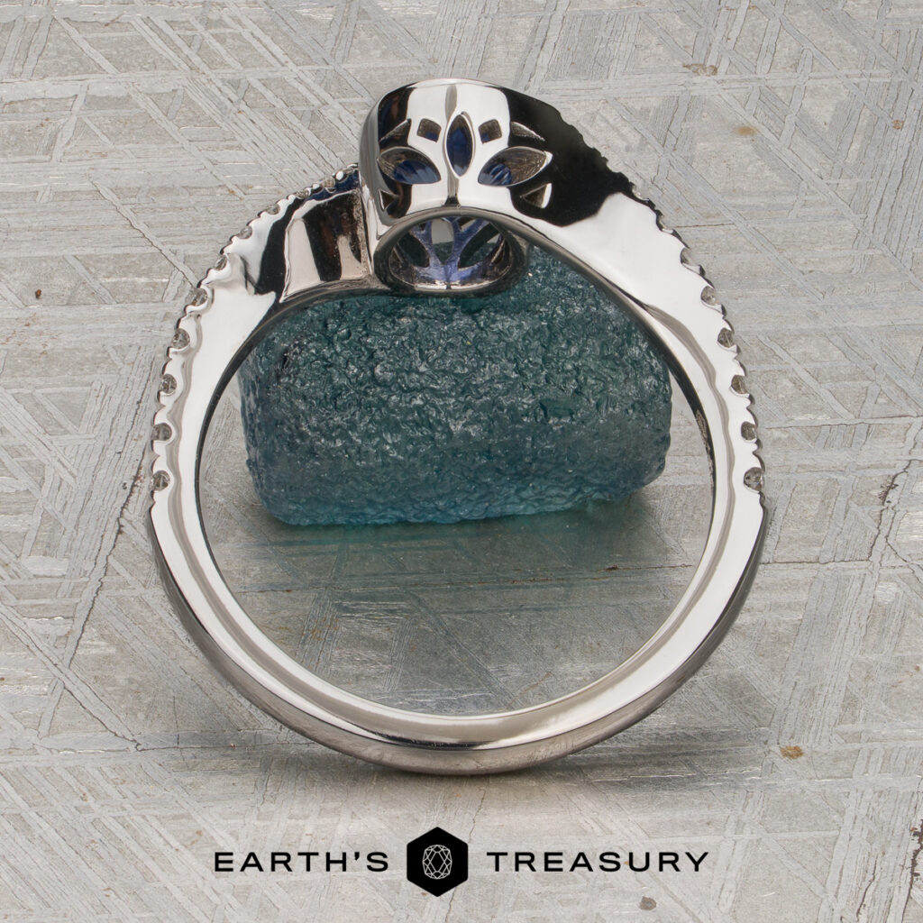 A textured silver ring, "Potiker, Cara," featuring a blue gemstone, rests against a rough blue crystal on a patterned surface adorned with the "EARTH'S TREASURY" logo. Intricate filigree details highlight the band, bringing an elegant touch to this distinctive piece.