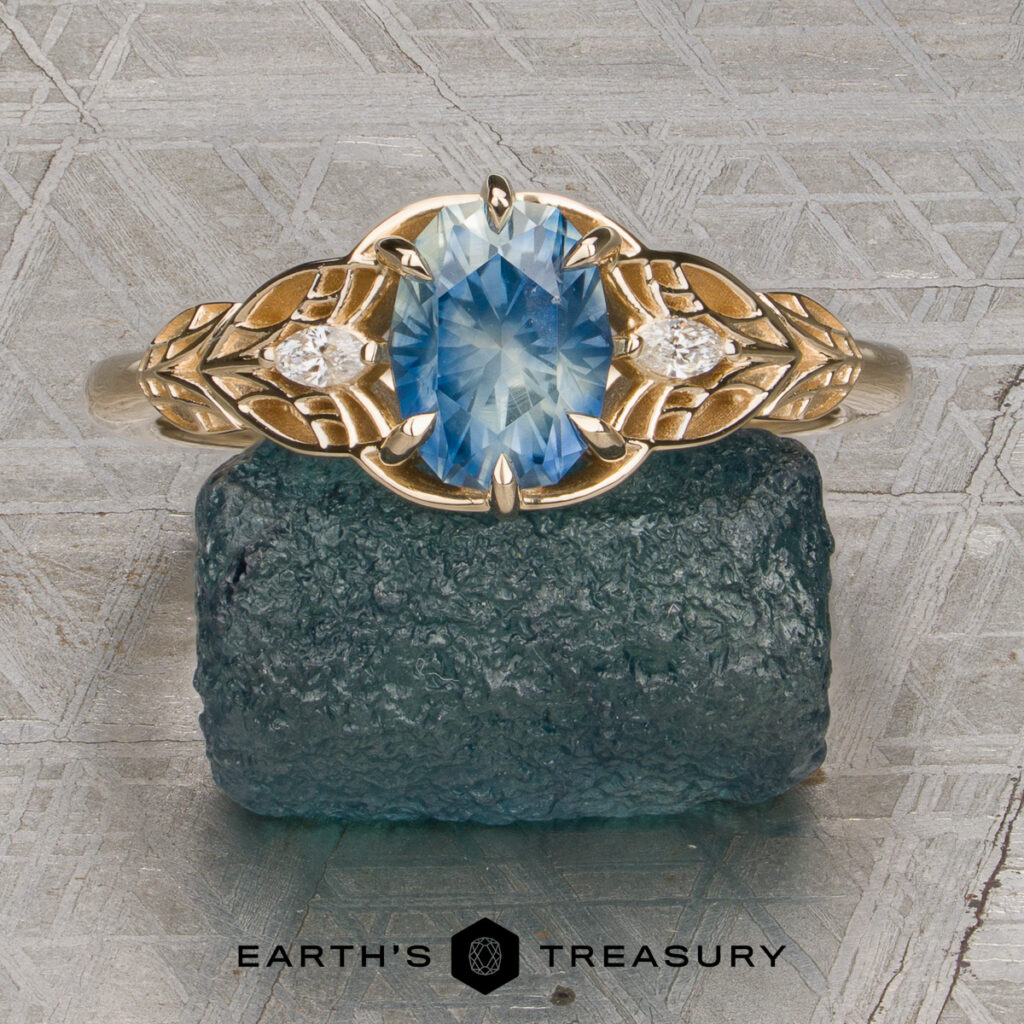A gold ring from the Thomas, Harrison [custom gallery] collection, featuring an oval blue gemstone and two small diamonds, rests on a rough blue stone. The background displays a patterned metallic surface with the "Earth's Treasury" logo at the bottom.