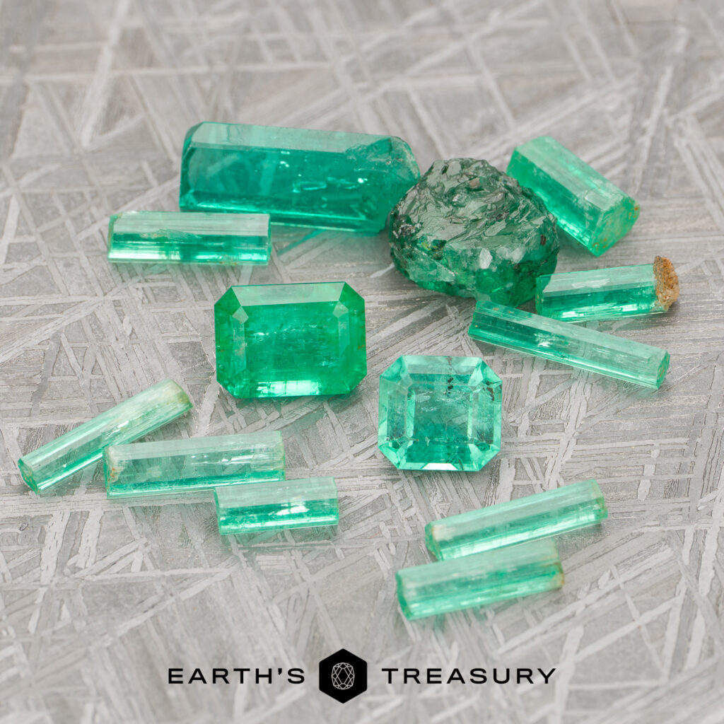 A collection of green emerald crystals and gemstones, both rough and faceted, are arranged on a textured gray surface. Captured in the product "Stacked from 8 images. Method=B (R=7,S=2)," the natural beauty of these gems shines through—reminiscent of features highlighted in any Emerald Guide—with the "Earth's Treasury" logo displayed at the bottom of the image.