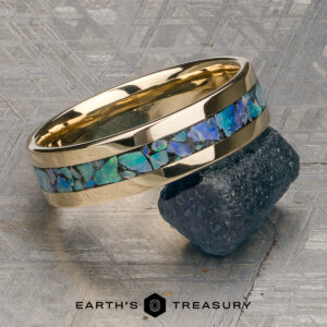 A Ready to Ship gold ring, named "Stacked from 50 images. Method=B (R=7,S=2)," featuring a blue-green opal inlay, is displayed resting on a dark gemstone against a geometric metallic background, with the Earth’s Treasury logo visible below. This elegant 14K Yellow Gold Triton Band exemplifies modern design and timeless craftsmanship.